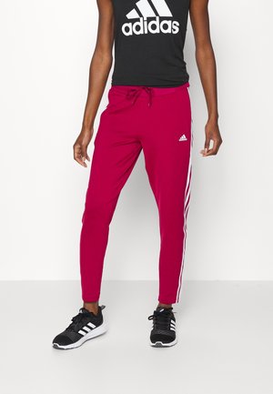 Rote Sport-Jogginghosen mit weißen Seitenstreifen und elastischem Taillenbund; kombiniert mit einem schwarzen Tanktop mit weißem Branding, getragen mit schwarzen Sneakers.