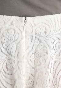 Weißer Spitzenstoff mit kunstvollen, wirbelnden Mustern und einem verdeckten Reißverschluss am Rücken. Der graue Hintergrund hebt die Struktur der Spitze hervor.