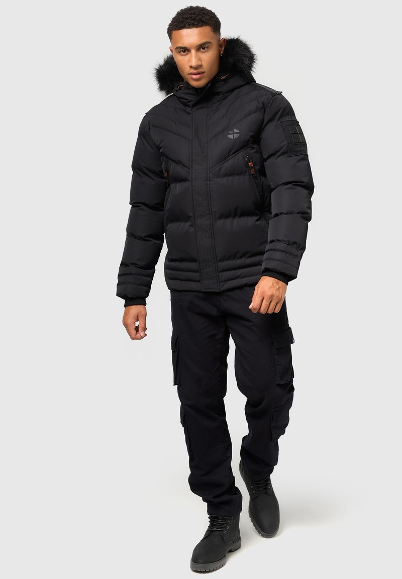 Svart puffjacka med en foderhuva av päls, dragkedjefickor och ribbade ärmar, tillsammans med svarta cargobyxor och snörstövlar.