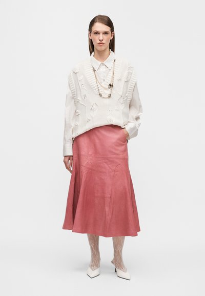 Gilet blanc en maille avec des motifs floraux texturés sur une chemise claire, associé à une jupe en cuir rose fluide et des chaussures blanches à bouts pointus.