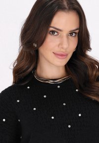 Schwarzer strukturierter Pullover mit Kristallverzierungen. Verfügt über eine metallische mehrsträngige Halskette. Warmtoniger, weicher Stoff mit unterschiedlicher Tiefe.