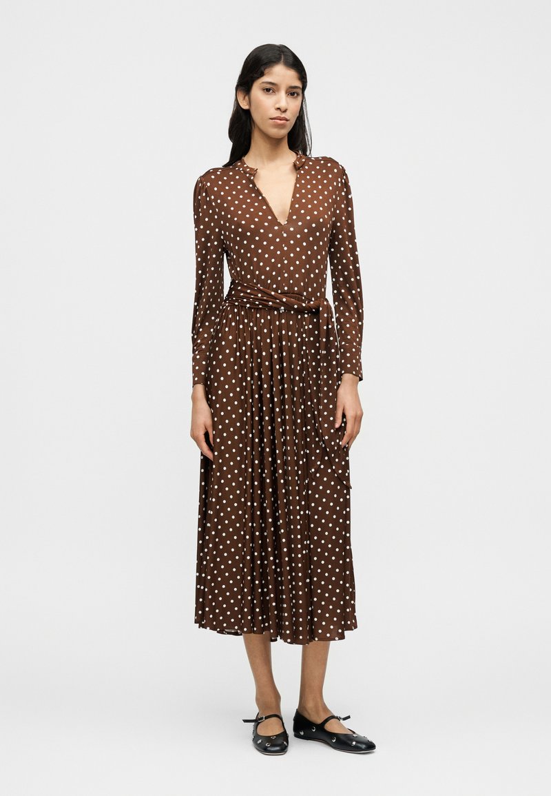 Καφέ φόρεμα με πουά, μακρυά μανίκια, V-λαιμόκοψη και ζώνη στη μέση. Λείο ύφασμα με φαρδιά φούστα, συνδυασμένο με μαύρα φλατ παπούτσια.