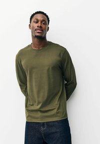 Nuori mies seisoo valkoisen seinän edessä yllään oliivinvihreä pitkähihaisen paita ja tummansiniset farkut, kädet selän takana, katsoo kameraan.