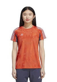 T-shirt de sport rouge avec un motif abstrait, manches courtes, accents bleus, et trois bandes blanches sur les épaules. Logo Adidas sur la poitrine.