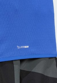 Sininen urheilupaita, jossa on teksturoitu kangas ja "CLIMAWARM" -merkintä helman kohdalla, yhdistettynä kuviollisiin mustiin shortseihin.