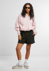 Lys rosa hettejakke med glidelås og frontlomme, kombinert med svarte shorts med et dekorativt mønster. Grå joggesko og hvite ankelsokker fullfører antrekket.