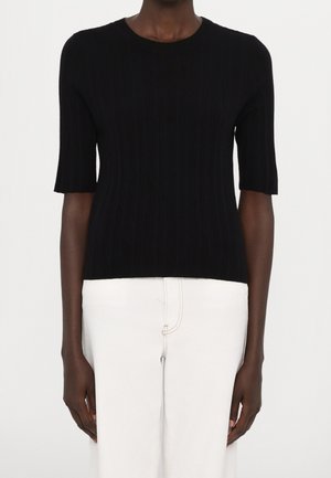 Donkere persoon die een zwart geribd truitje met korte mouwen en een witte broek met hoge taille draagt, staand tegen een effen achtergrond.