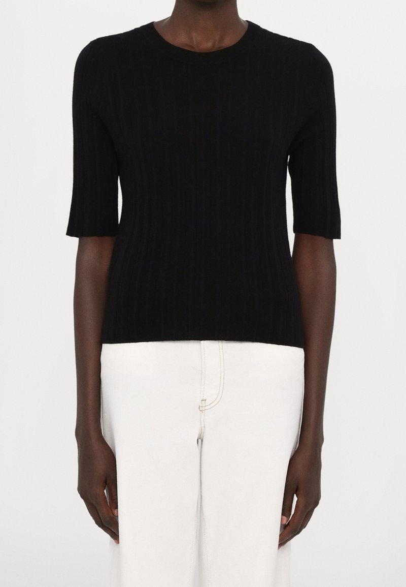 Personne à la peau foncée portant un pull noir à manches courtes côtelé et un pantalon blanc taille haute, debout devant un fond uni.