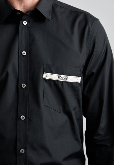 Cămașă neagră cu nasturi, cu un design elegant, având un buzunar pe piept cu un accent în dungi albe și eticheta "MOSCHINO". Textură netedă.