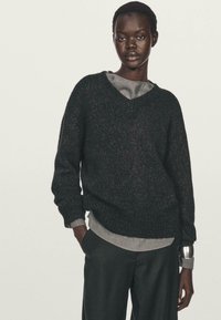 Svart stickad tröja med V-ringning, ribbade muddar och nederkant. Bärs över en grå långärmad top. Modellen står med händerna i fickorna, iförd mörka byxor.