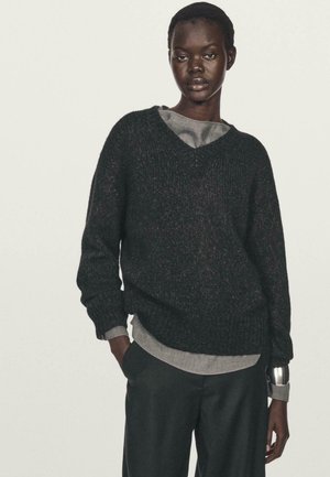 Sort strikket sweater med V-hals, ribbestrikkede manchetter og kant. Bæres over en grå langærmet top. Modellen står med hænderne i lommerne og har mørke bukser på.