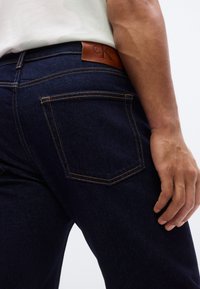 Σκούρο μπλε τζιν με υφή. Περιλαμβάνει αντίθετη πορτοκαλί ραφή και καφέ δερμάτινο ταμπελάκι στη μέση.