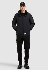 Mężczyzna w czarnej kurtce bomberze z futrzanym kołnierzem, czarnych spodniach cargo, czarnych butach i czapce z daszkiem, stoi z dłońmi w kieszeniach.