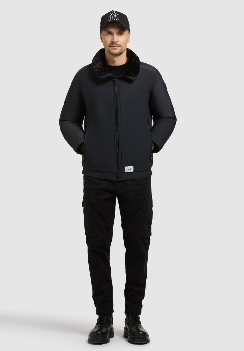 Mężczyzna w czarnej kurtce bomberze z futrzanym kołnierzem, czarnych spodniach cargo, czarnych butach i czapce z daszkiem, stoi z dłońmi w kieszeniach.