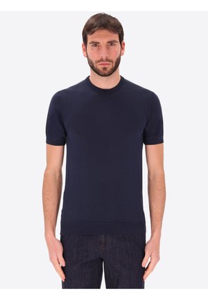 Uomo con capelli scuri e barba che indossa un maglione semplice blu navy a maniche corte e collo tondo e jeans scuri, in piedi davanti a uno sfondo chiaro.