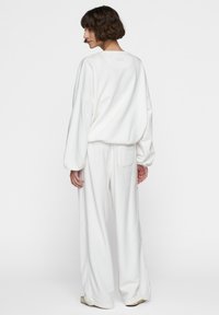 Donna con capelli corti e ricci, che indossa un lungo top bianco a maniche larghe e pantaloni larghi, in piedi e che guarda oltre la spalla su uno sfondo bianco.