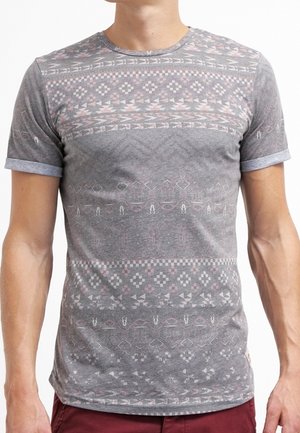 Mężczyzna noszący dopasowany szary T-shirt z subtelnymi, geometrycznymi i plemiennymi wzorami w jasnoróżowym i białym kolorze, zestawiony z bordowymi spodniami.