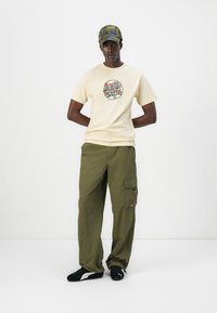 Short yellow t-shirt with floral graphic, paired with loose olive green cargo pants and black athletic shoes featuring white accents.