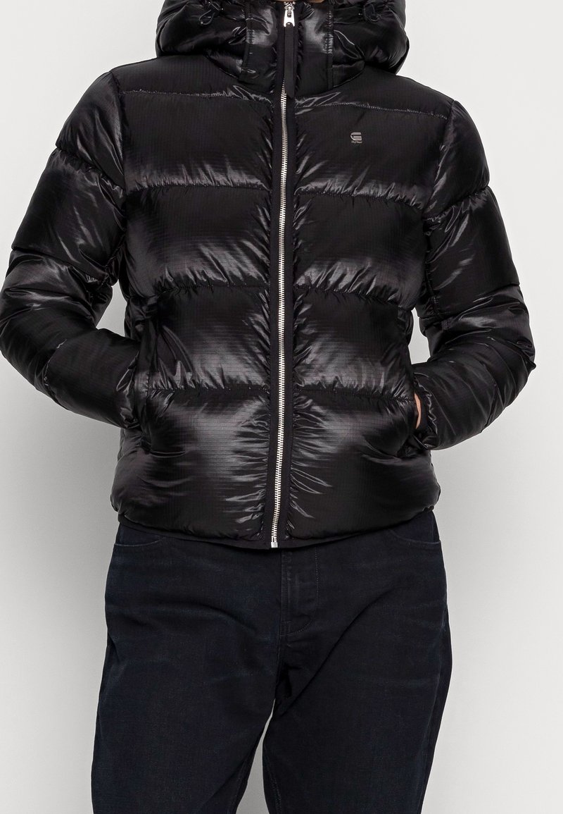 Personne portant une doudoune noire brillante avec les mains dans les poches, une fermeture éclair frontale et un petit logo sur la poitrine, associée à un pantalon foncé.