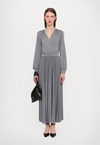 Robe grise à manches longues avec un design enroulé, jupe plissée et taille cintrée. Accessoirisée avec un sac noir et des escarpins à bout pointu.