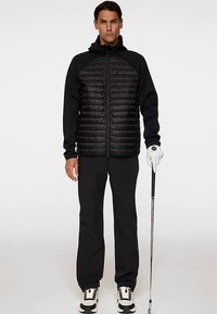 Svart quiltad jacka med luva, ärmar i kontrasterande slät tyg, kombinerad med svarta byxor och golfhandske, håller en golfklubba.