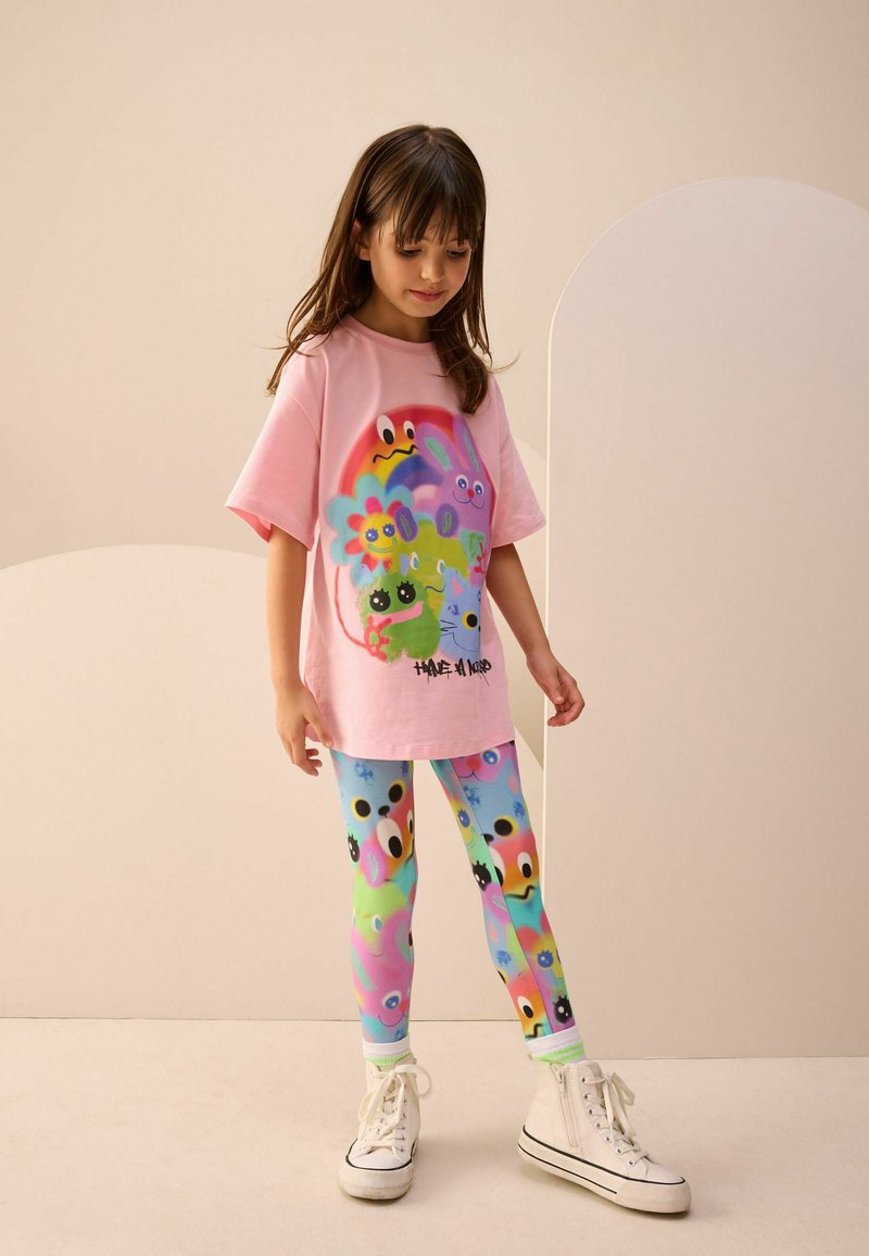 Rosa Baumwoll-T-Shirt mit bunten Cartoon-Grafiken, kombiniert mit gemusterten Leggings in Blau- und Grüntönen, die niedliche Charakterdesigns zeigen.