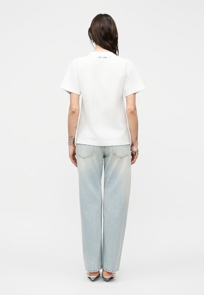 Άτομο με μακριά σκούρα μαλλιά που φοράει λευκό μπλουζάκι με κοντά μανίκια, ανοιχτό μπλε τζιν παντελόνι με φαρδιά μπατζάκια και παπούτσια με τακούνι, όρθιο με την πλάτη γυρισμένη.