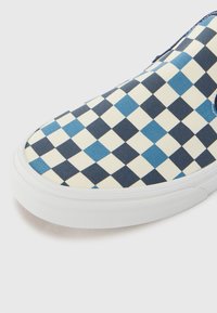 Zapatos slip-on de cuadros azules y crema con parte superior de lona, suela de goma y ribete texturizado blanco, destacando un patrón de cuadros contrastante.