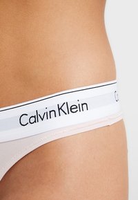Calvin Klein tangaslip in lichtbeige stof, met een gladde textuur en een witte elastische tailleband met zwart merkembleem.