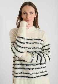 Maglione a maglia crema con righe orizzontali nere, dotato di colletto alto e polsini a coste. Texture morbida, vestibilità rilassata con fessure laterali.
