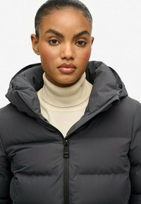 Mørkegrå dunjakke med lynlås og en stor, polstret hætte. Båret over en lys beige turtleneck. Glat tekstur, quiltet design.