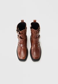 Bruna läderankelboots med tre silverfärgade spännen och svarta sulor, fotograferade ovanifrån mot en vit bakgrund.