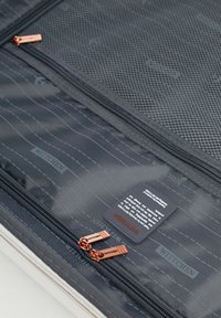 Interior de la maleta con tejido oscuro, un bolsillo de malla, dos cremalleras en color rosa dorado y la marca WITCHEH en una etiqueta y patrón grises.