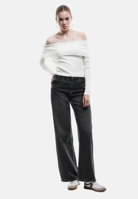 Jonge vrouw in een witte off-shoulder ribgebreide trui, brede zwarte jeans, witte sneakers en grote ringoorbellen tegen een effen achtergrond.