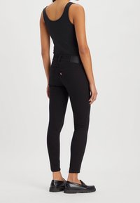 Black skinny jeans with a high waist, featuring a leather back patch and subtle logo detail. Paired with a black tank top and black shoes.