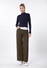 Marineblå ribbestrikket genser med glidelås over en hvit skjorte, kombinert med vide olivengrønne bukser og hvite joggesko med svarte striper.