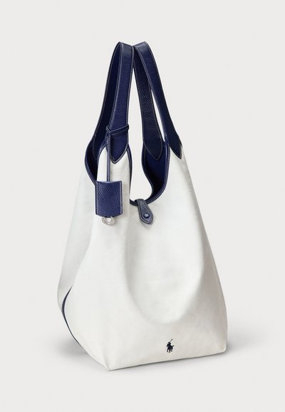 Λευκή καμβά τσάντα tote με σκούρα μπλε δερμάτινες λαβές και διακόσμηση, με ένα μικρό σκούρο λογότυπο κοντά στη βάση και μια δερμάτινη ετικέτα σε μία από τις λαβές.