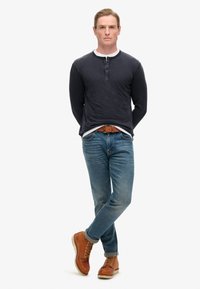 Dunkelblaues Langarm-Henley-Shirt mit weißem Kragen, kombiniert mit hellblauen Jeans und braunen Schnürstiefeln. Das Model steht in einer entspannten Pose.