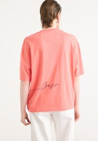 Coral cotton t-shirt with short sleeves, a round neckline, and a handwritten "Boss" graphic in black on the back. Flat texture.