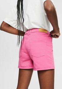 Vaaleanpunaiset puuvillashortsit klassisella leikkauksella, joissa on kaksi takataskua ja ruskea nahkapatch vyötäröllä. Strukturoidut materiaalit.