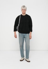 Svart fleece sweatshirt, ljusblå denimjeans och beige sneakers. En grå axelremsväska med justerbar rem. Enfärgad bakgrund.