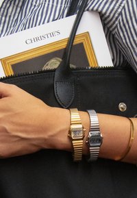 Twee digitale horloges met rechthoekige wijzerplaten: één goudkleurig met een metalen band, de ander zilverkleurig. Armband met een minimalistisch ontwerp; hand die een zwarte tas vasthoudt.