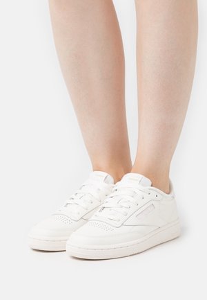 White low-top sneakers with laces worn on bare feet, visible stitching, and subtle brand logo on a plain white background.