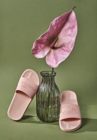 Różowe gumowe klapki z teksturowanymi podeszwami i wytłoczonym logo, zestawione z przezroczystym szklanym wazonem zawierającym dużą różową roślinę, na zielonym tle.