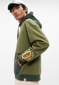 Ελαιοπράσινη φούτερ με κουκούλα και μεγάλη τσέπη, με κεντητό σχέδιο στο μανίκι που απεικονίζει μια ουράνια θεματολογία. Υλικό μείγμα βαμβακιού.