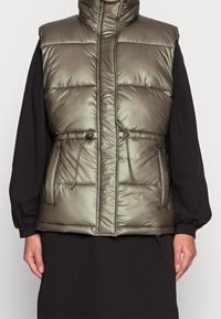 Khaki kamizelka puchowa z wysokim kołnierzem, bocznymi kieszeniami i regulowanym sznurkiem w talii. Błyszcząca, pikowana struktura o lekkiej konstrukcji.