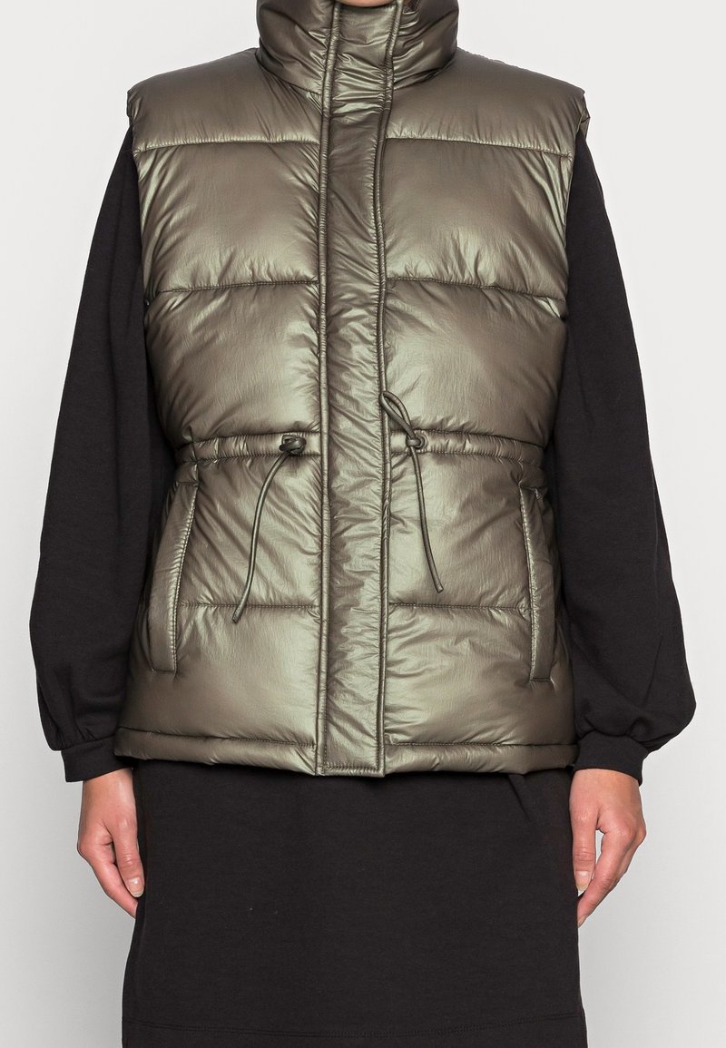 Khaki kamizelka puchowa z wysokim kołnierzem, bocznymi kieszeniami i regulowanym sznurkiem w talii. Błyszcząca, pikowana struktura o lekkiej konstrukcji.