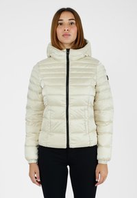 Giacca cremosa imbottita con cappuccio, dotata di zip nera, polsini elasticizzati e texture trapuntata. Indossata con pantaloni neri.