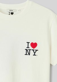 T-shirt en coton crème avec col rond, présentant un « I » et « NY » noirs, ainsi qu'un cœur rouge imprimé sur le côté gauche de la poitrine.