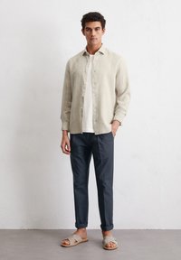 Chemise en lin beige clair à boutons sur un t-shirt blanc, associée à un pantalon bleu marine et des sandales grises, se tenant contre un fond neutre.